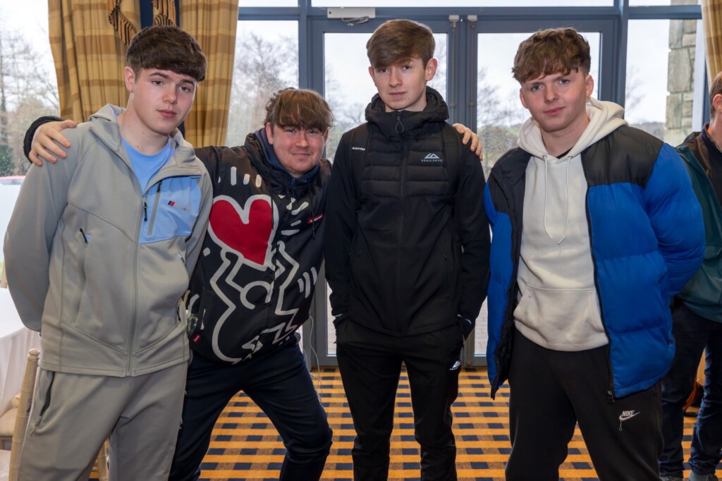 Four young males stand side by side indoors with their arms around each other.