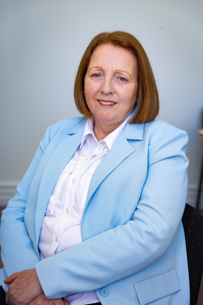 Photo of lady in light blue blazer and white blouse, sitting and smiling.